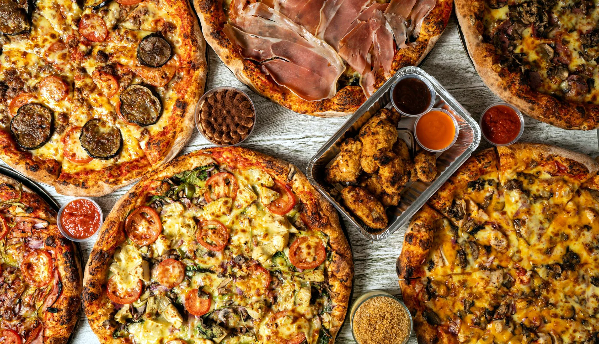 Overhead spread of specialty pizzas with chicken wings and dips